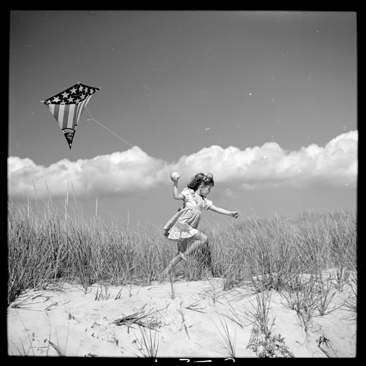 Photograph shows Toni Frissell's daughter, Sidney Bacon (Frissell) Stafford, running on a beach with a kite in Southampton, Long Island, New York in 1944 - by Toni Frissell.